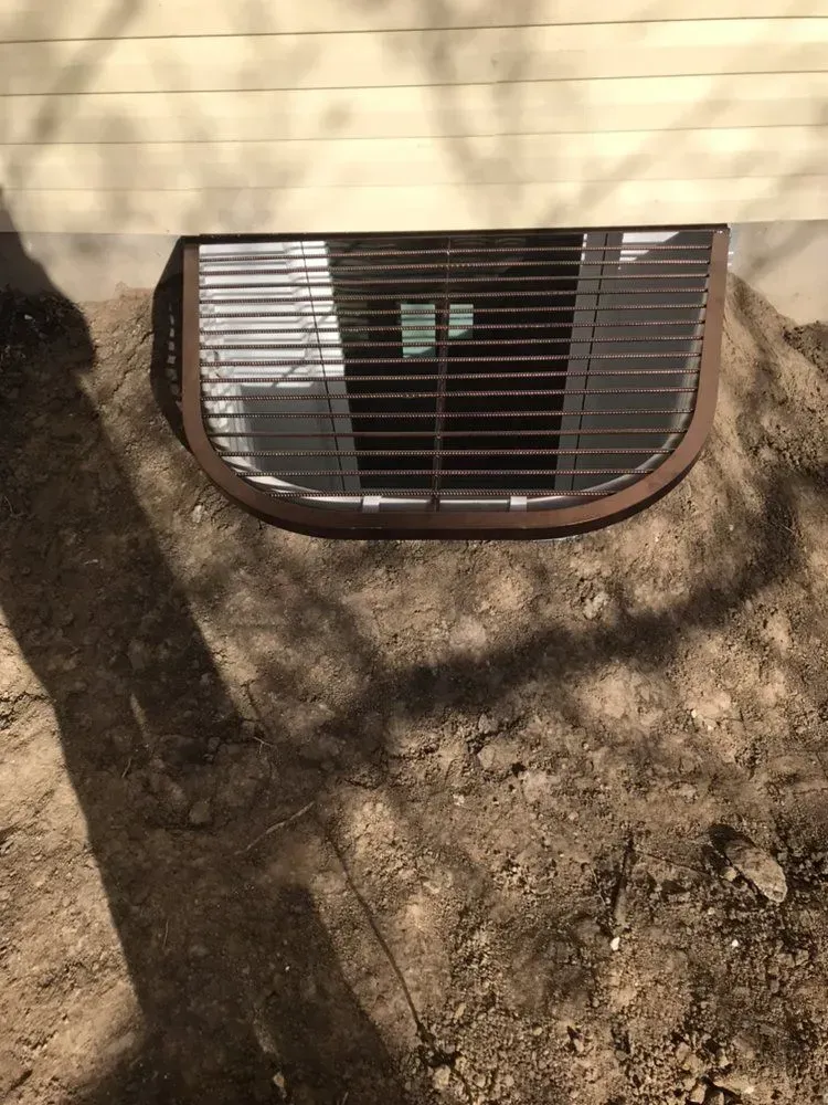 Exterior basement window well with a black metal grate in a dirt embankment