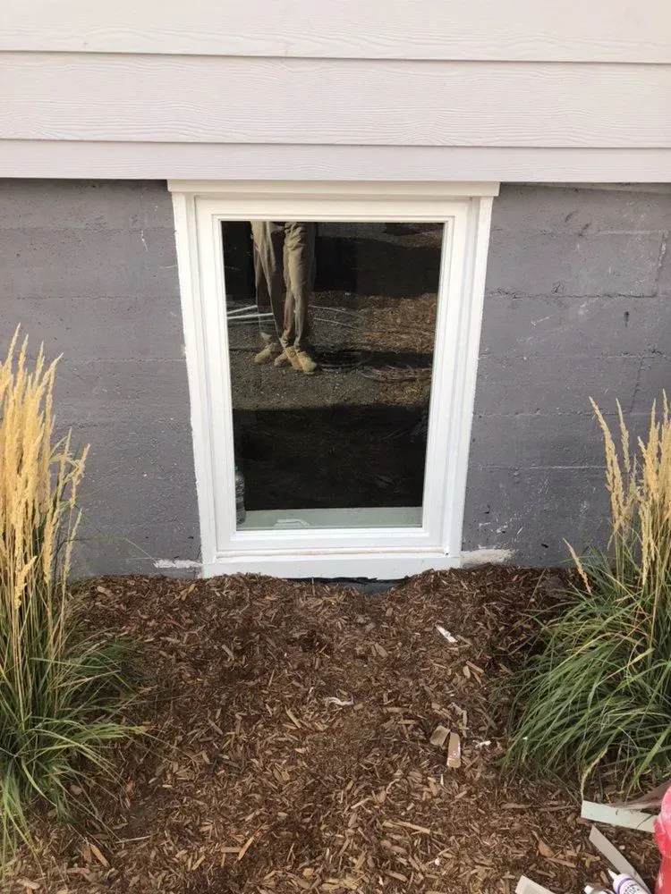 Basement window well with white trim beneath gray siding, surrounded by mulch and landscaping.