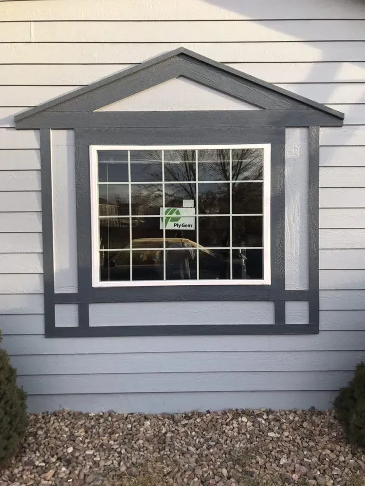 Gray house window with dark trim and a small sign, set in light siding above gravel.