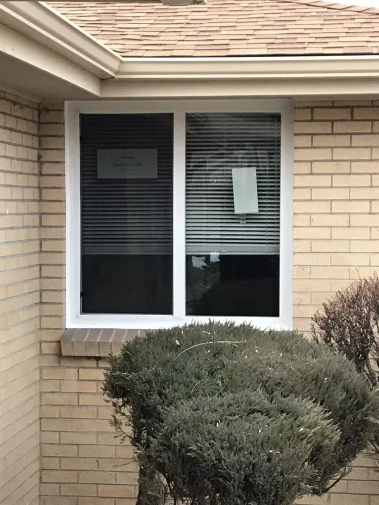 House window with white blinds and a posted notice above shrubs on a brick exterior