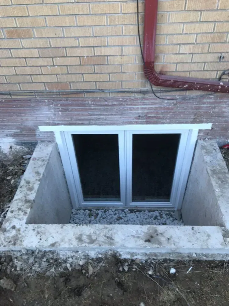 Basement window well with two sliding windows beneath a brick wall and red pipe