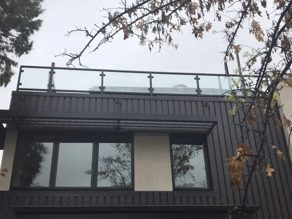 Modern dark-gray house facade with large windows and rooftop glass railing, framed by tree branches