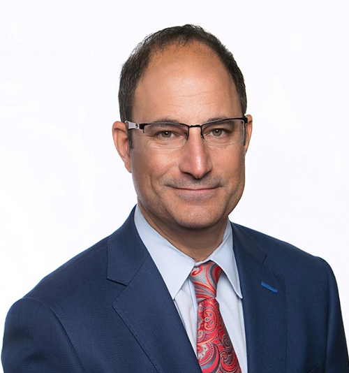 A man in a blue suit and white shirt is smiling in front of a brick wall.