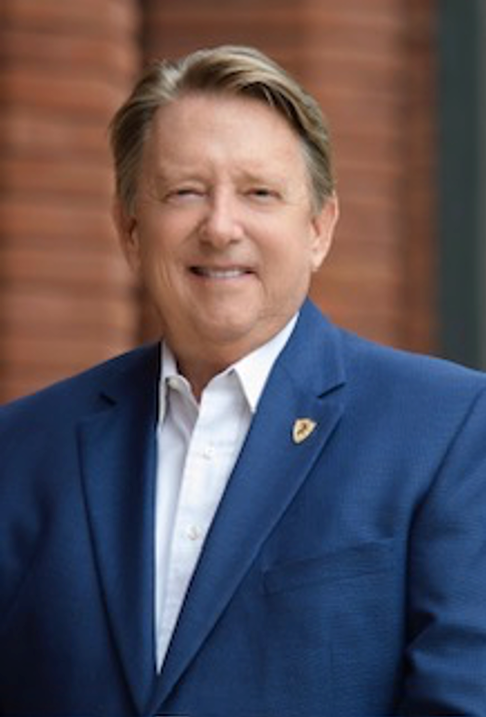A man in a blue suit and white shirt is smiling in front of a brick wall.