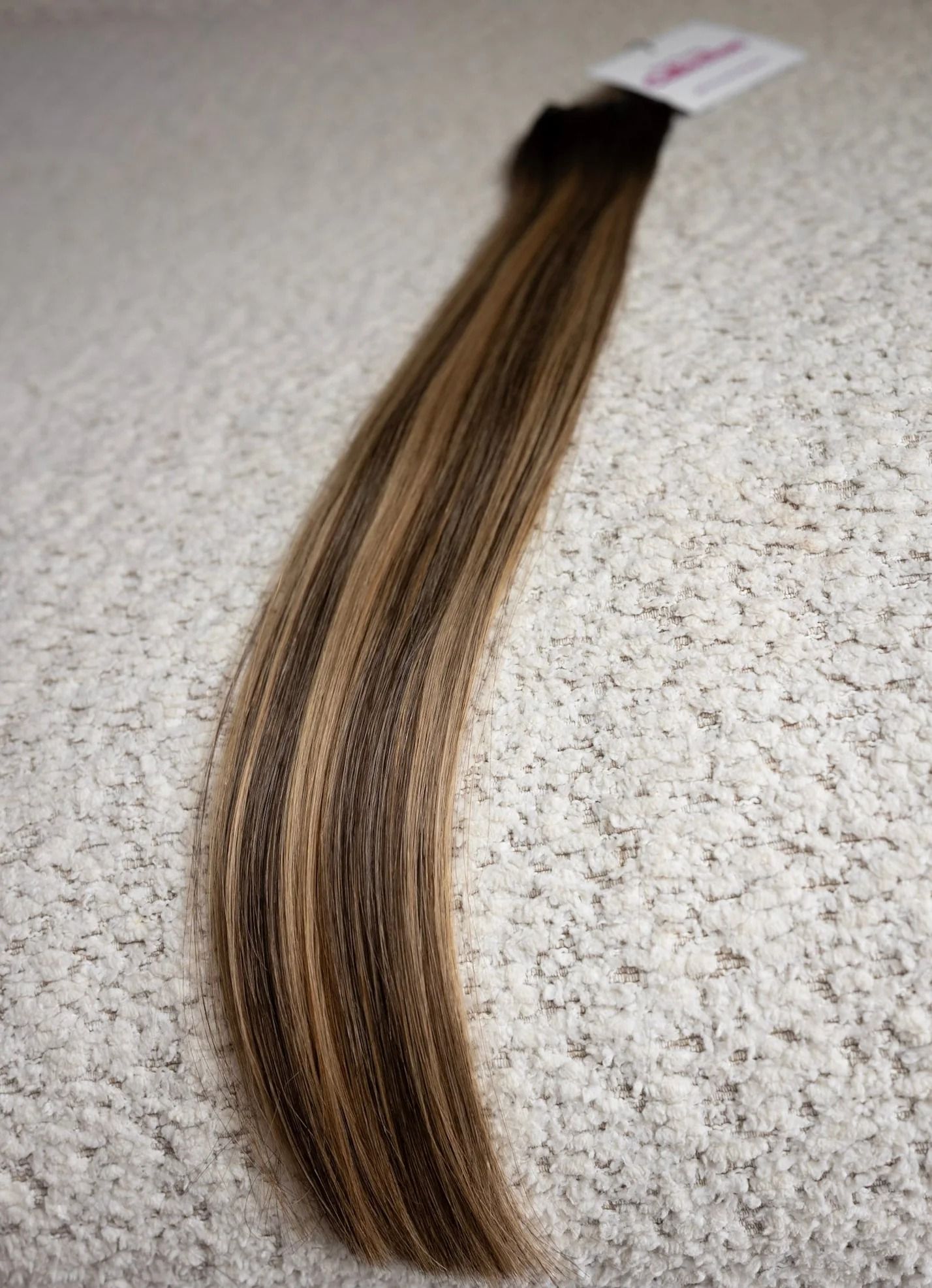 Long brown hair extension laid on a speckled carpet, with a small tag attached.