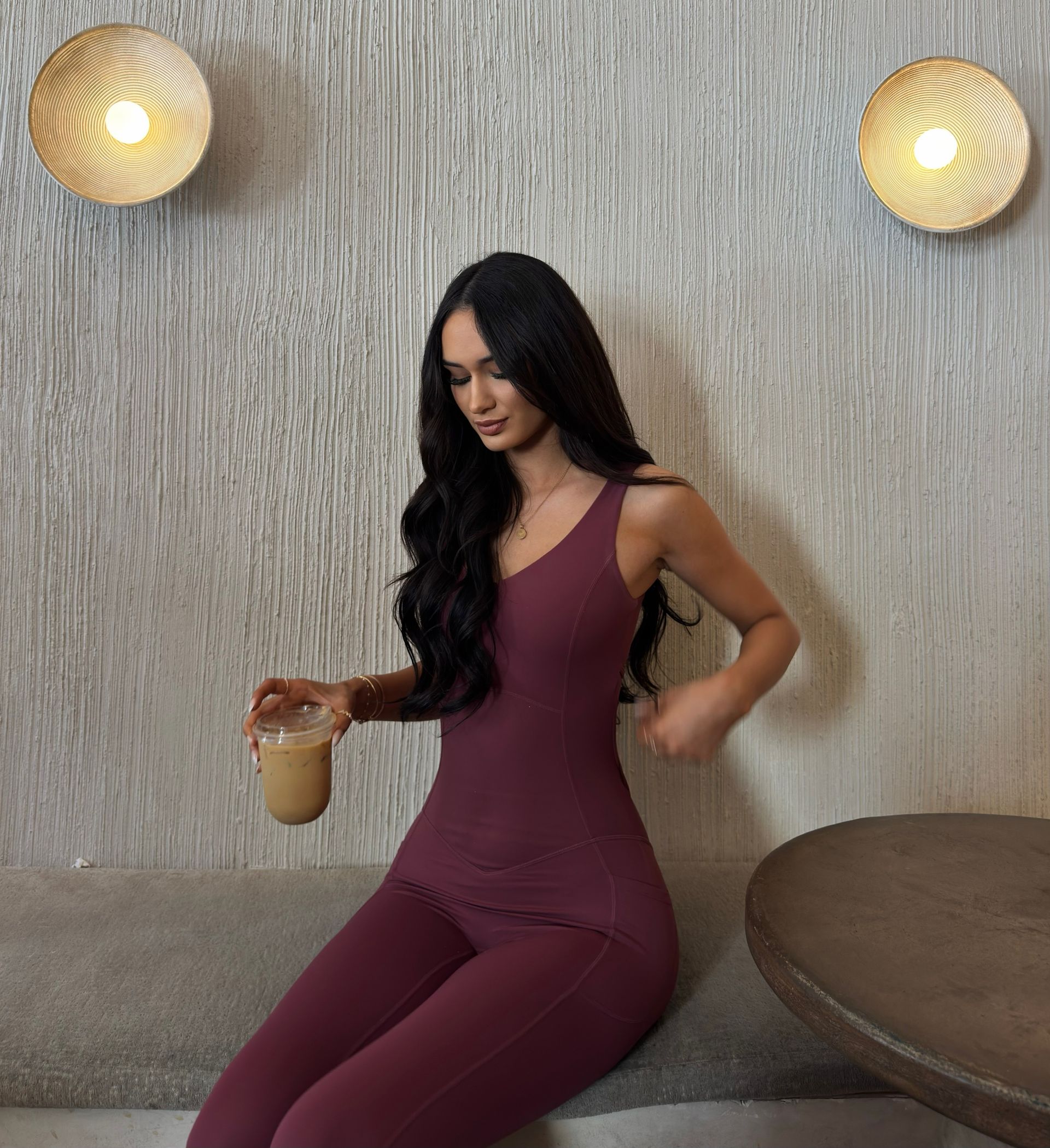 Woman in a burgundy jumpsuit holding a drink, seated in a cozy lounge with textured walls and warm wall lights.