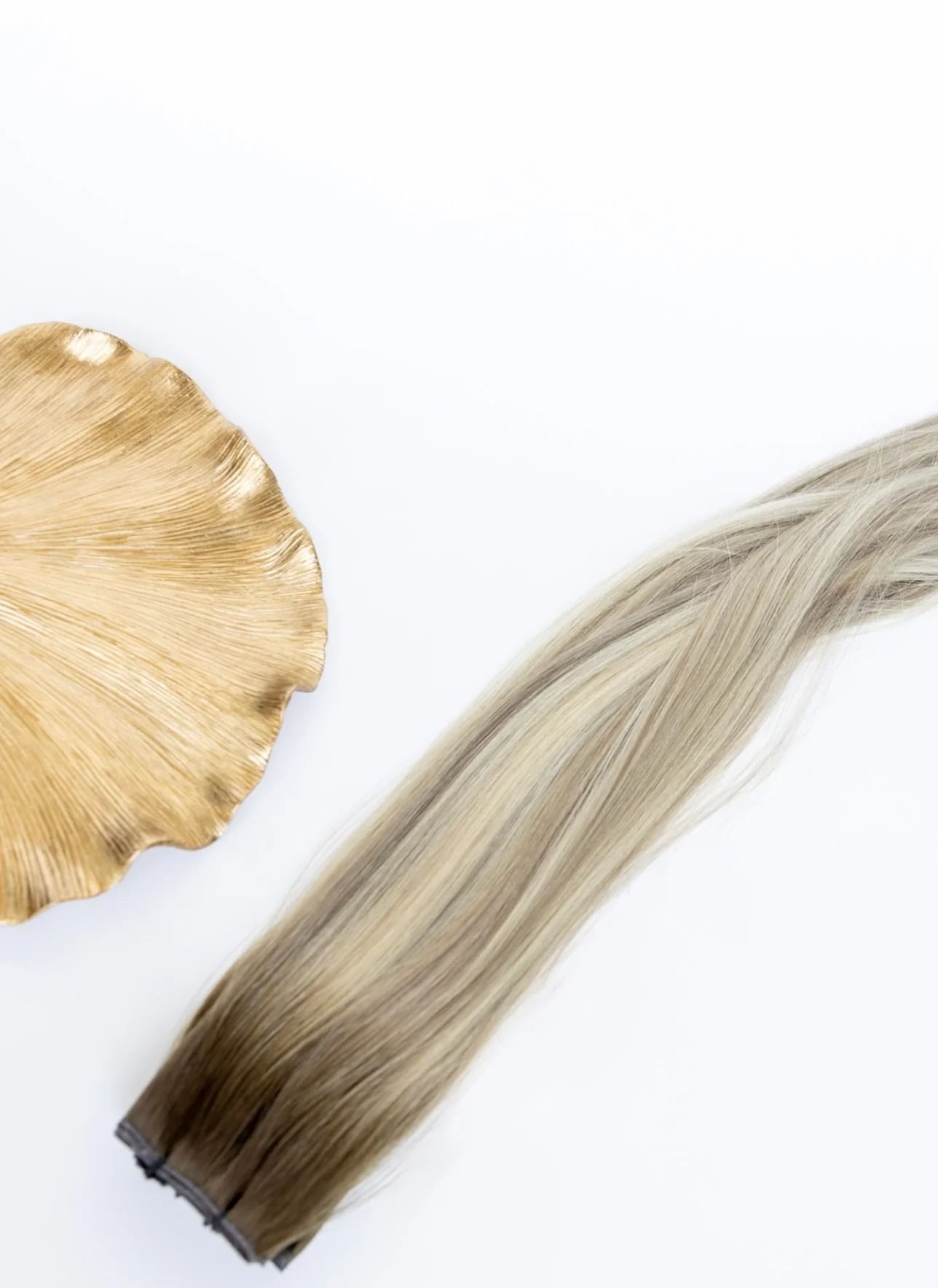 Blonde hair extension next to a gold-toned leaf-shaped hair accessory on a white background