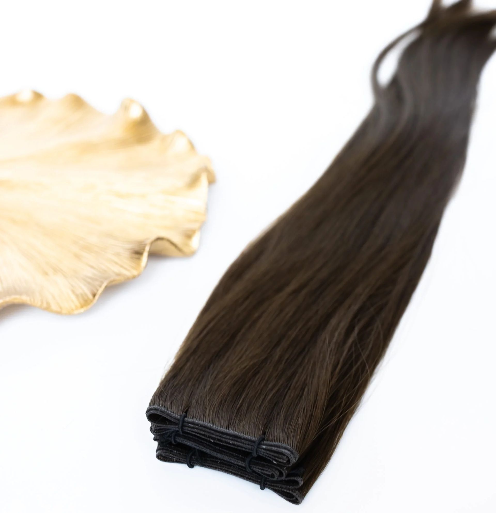 Dark brown hair extension bundle beside a cream seashell on a white background