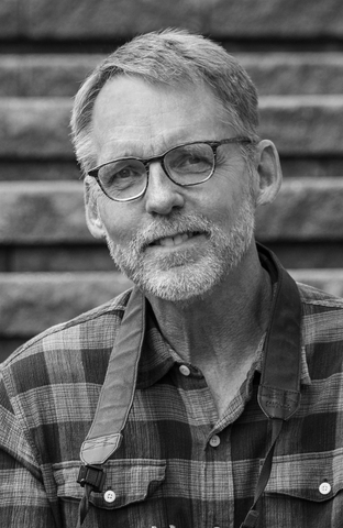 Portrait of a man with glasses and a gray beard, wearing a blue plaid shirt against a brick wall.