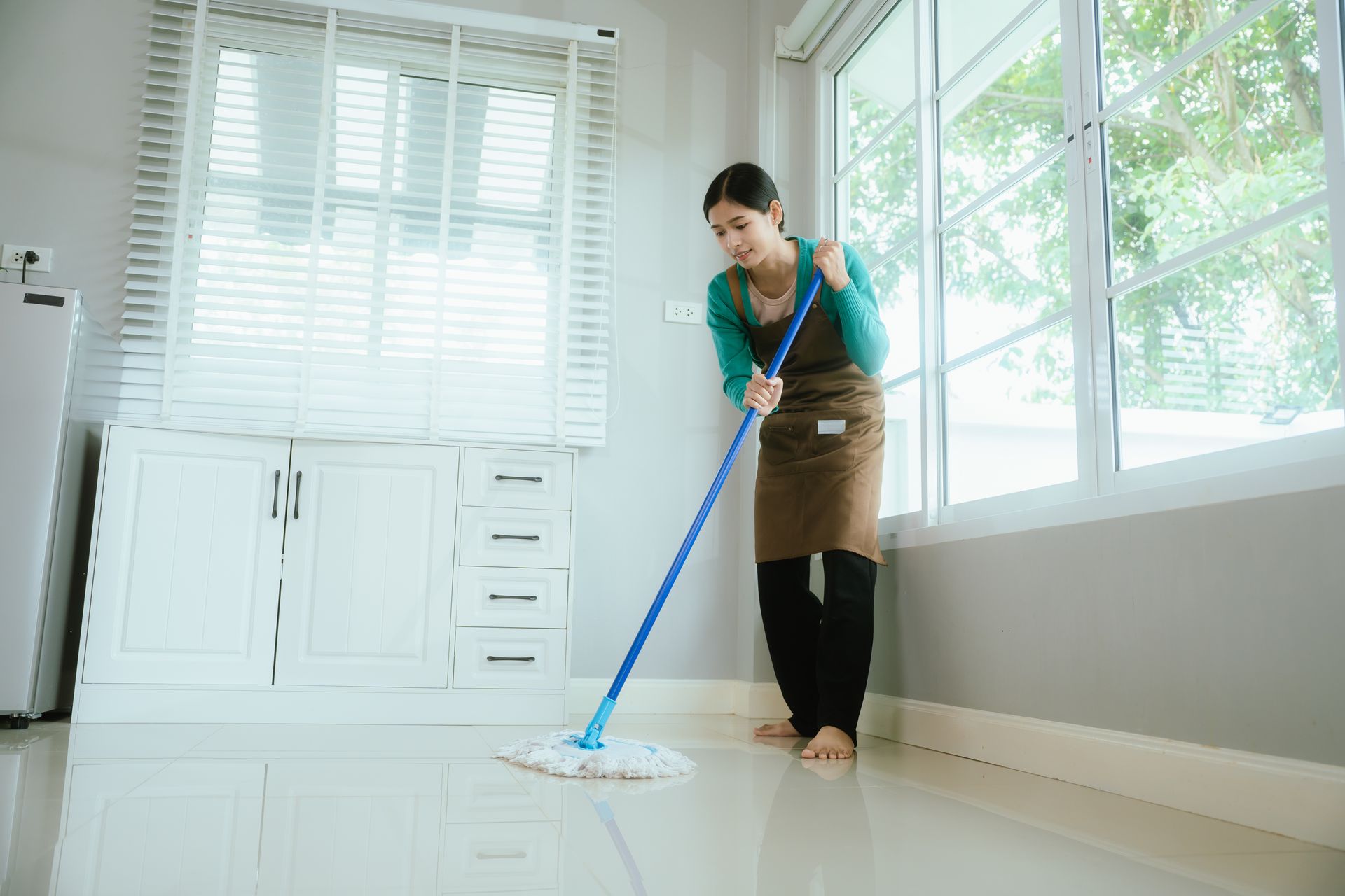 Person mopping a bright room with large windows and white cabinets Person mopping a bright room with large windows and white cabinets