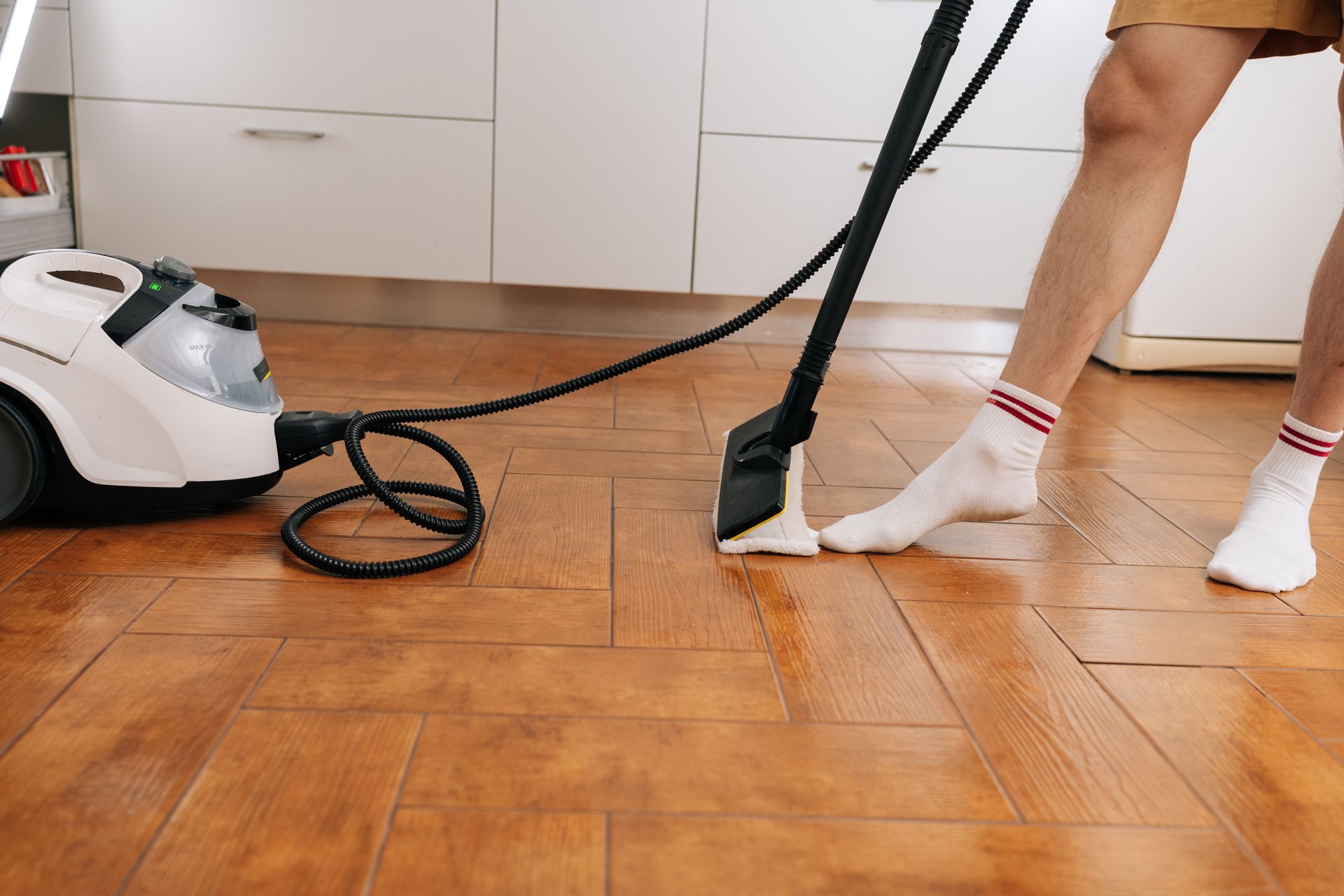 Person mopping a wooden kitchen floor with a white steam cleaner nearby Person mopping a wooden kitchen floor with a white steam cleaner nearby