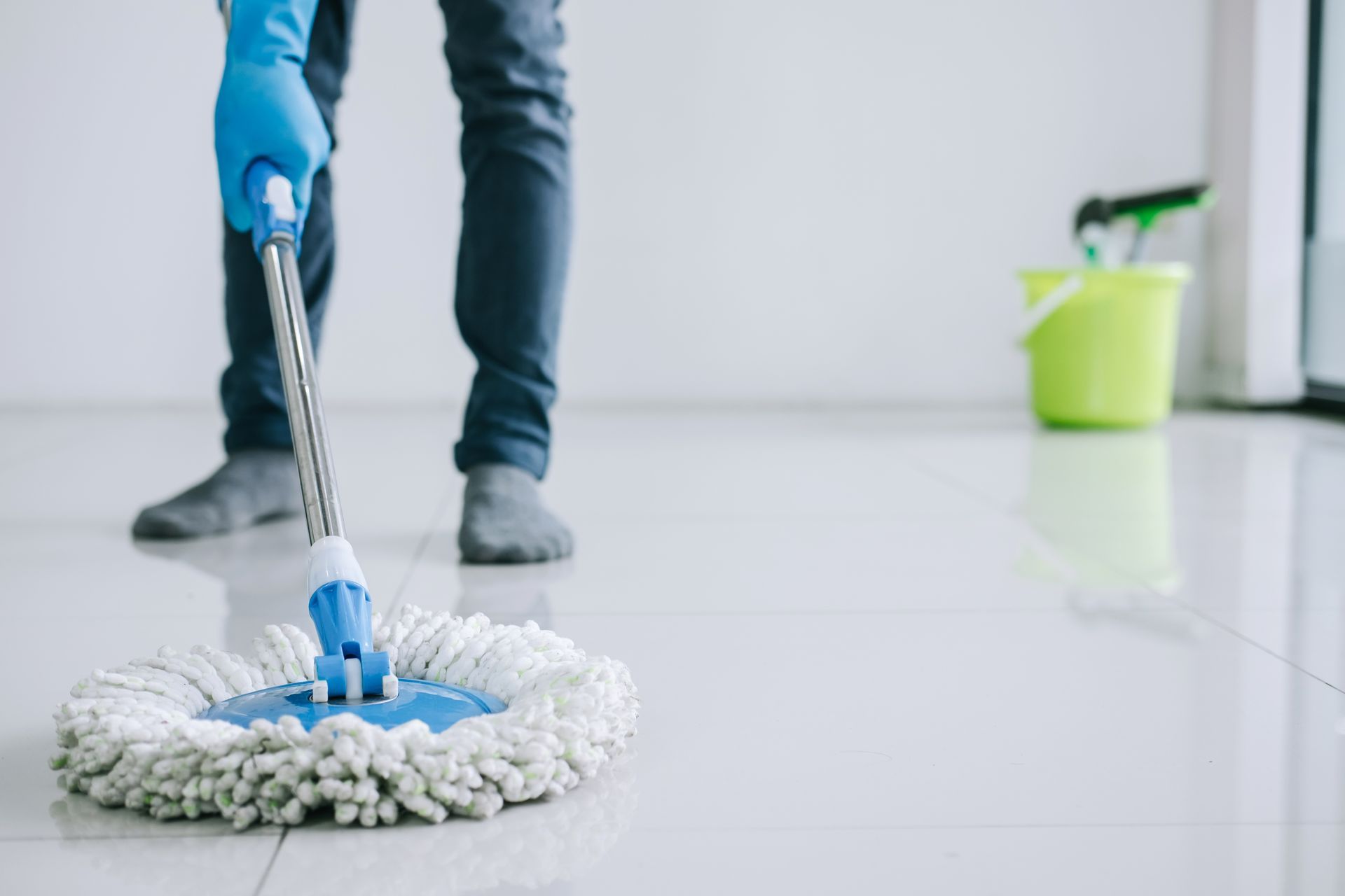 Person mopping a light-colored floor with a blue mop, wearing gloves and dark pants, with a bucket in the background. Person mopping a light-colored floor with a blue mop, wearing gloves and dark pants, with a bucket in the background.
