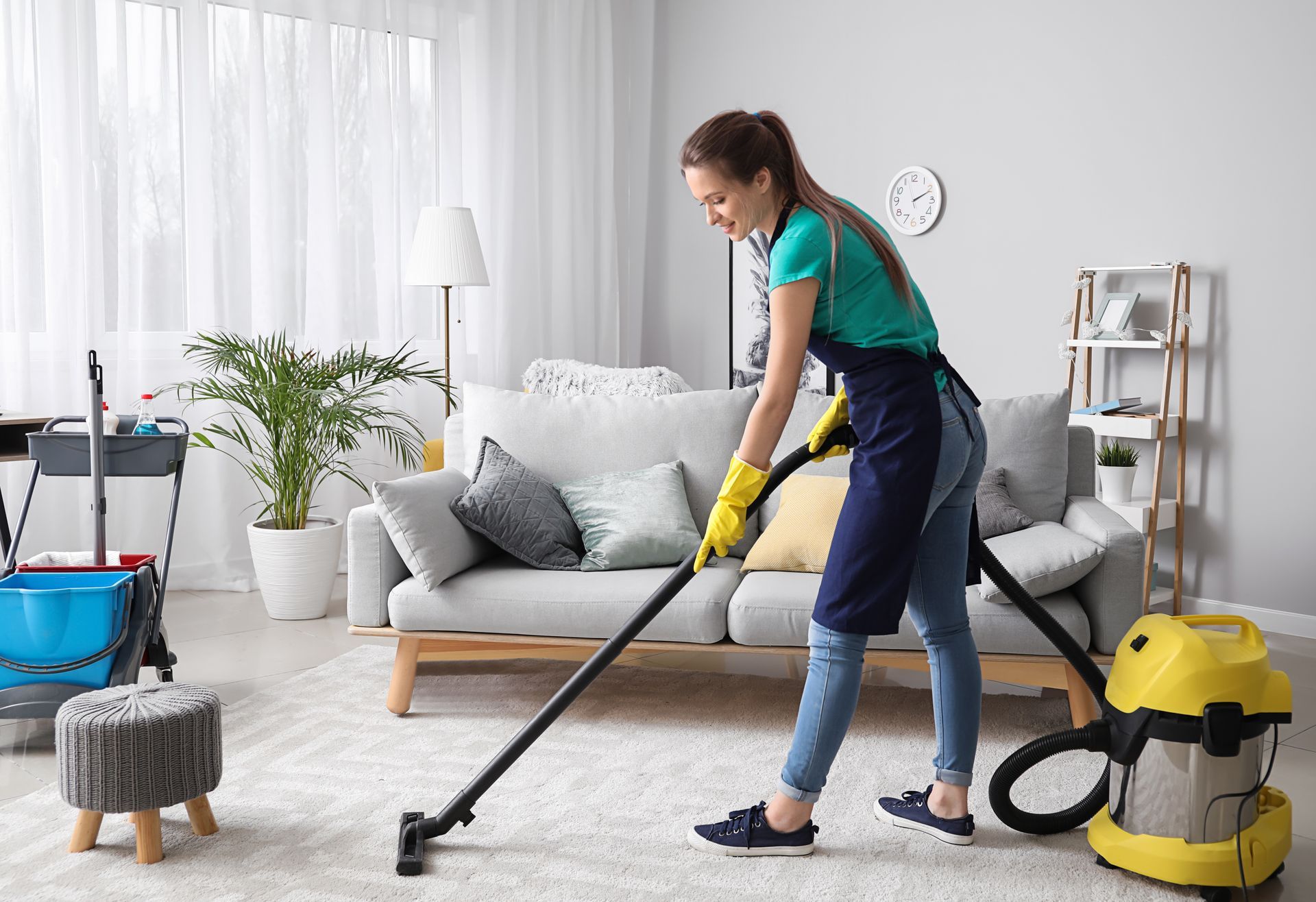 Woman vacuuming a carpet in a living room, wearing gloves and an apron, with cleaning supplies nearby.
