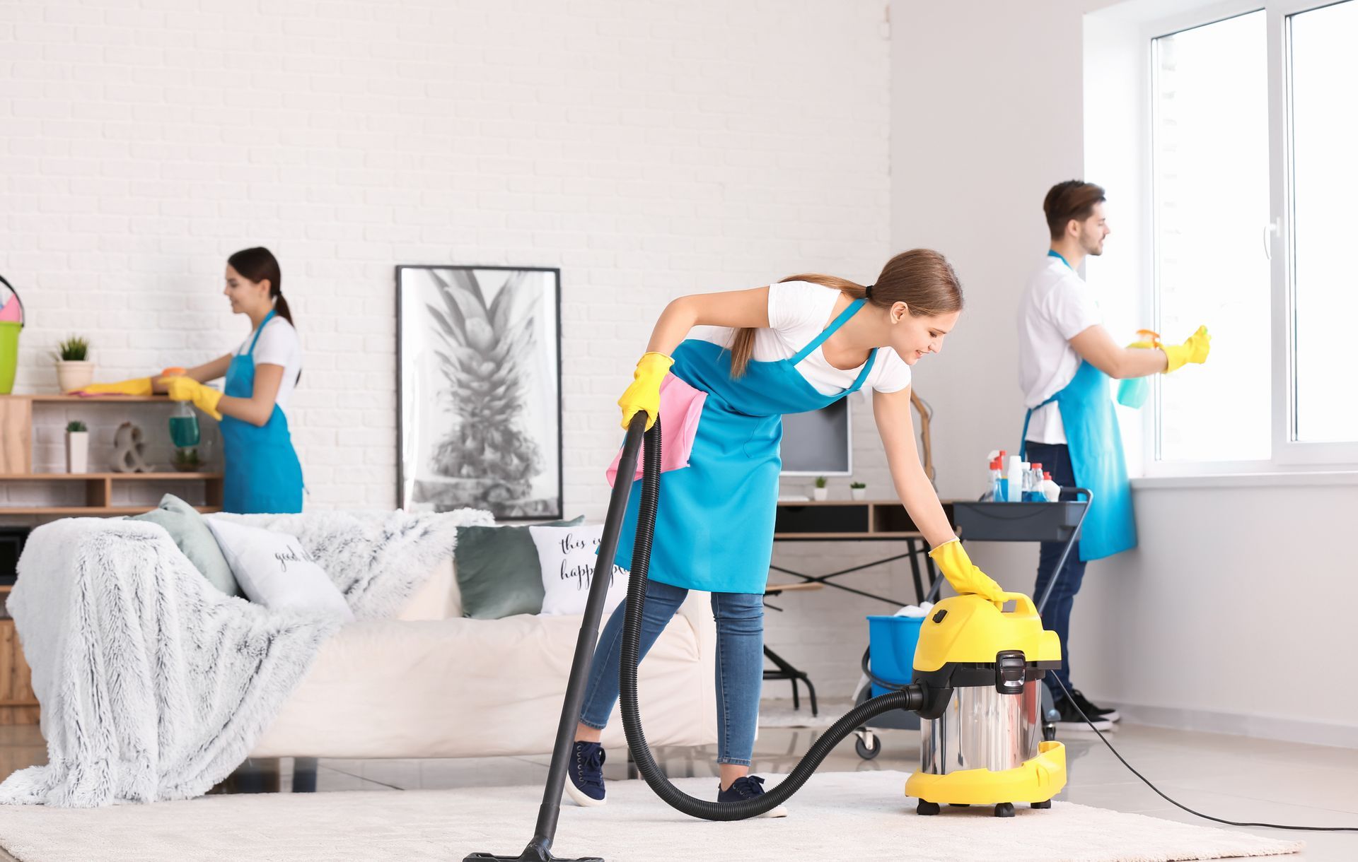 Three cleaners vacuum and dust in a bright living room with a sofa and large window. Three cleaners vacuum and dust in a bright living room with a sofa and large window.