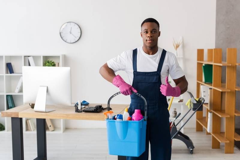 Woman vacuuming a light-colored rug in a living room; wearing an apron and gloves. Man in overalls, pink gloves, holding cleaning supplies bucket, thumbs up, office setting.