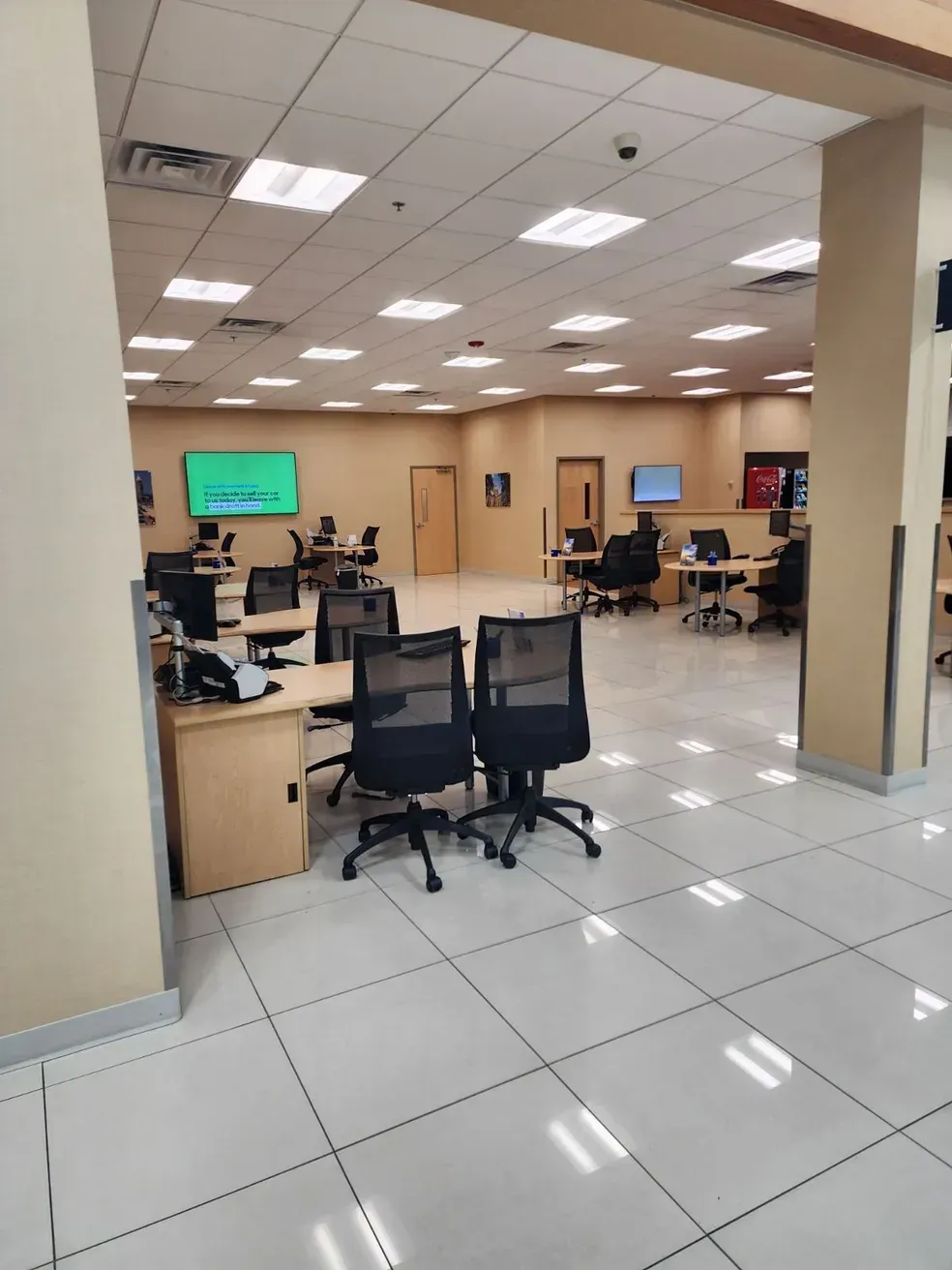 Interior of an office with desks, chairs, and monitors. Beige walls, white tiled floor, and overhead lighting. Interior of an office with desks, chairs, and monitors. Beige walls, white tiled floor, and overhead lighting.