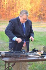 Man in dark suit adjusting a model cannon on a picnic table outdoors, with an American flag beside it.