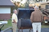 Two people standing beside a dark smoker grill outdoors near a house at dusk
