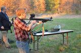 Person aiming a rifle at an outdoor shooting range, with another person standing nearby.