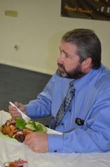 Man in a blue shirt and tie eating a meal at a table, holding a fork and looking to the side.