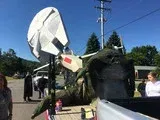 Person beside a large black sculpture holding a white, tilted satellite dish outdoors under a blue sky