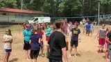 People gathered outdoors on a sandy field near a parked bus, watching an activity.