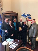 Group of people posing indoors with a framed award plaque, blue ribbon backdrop, and a desk in the foreground