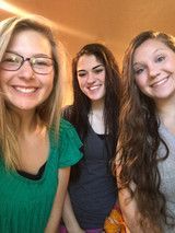 Three smiling people leaning close together indoors, with blonde, dark, and wavy brown hair.