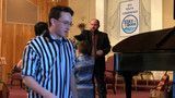 Man in striped shirt speaking beside a piano at a small indoor event