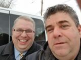 Two smiling people posing for a selfie in front of a white van outdoors