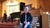 Man in a suit sits on a stage beside a sign in a church-like hall.