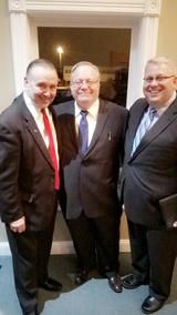 Three men in suits posing together indoors, smiling in a doorway.