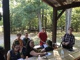 People sitting on a wooden deck in a wooded park, gathered around a table outdoors.