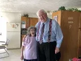Two people smiling indoors beside a wooden cabinet, with a refrigerator and folding chair in the background