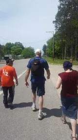 Three people walking on a sunny road with trees lining both sides