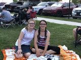 Two people sitting on an orange blanket at a grassy park, with parked cars and lawn chairs behind them