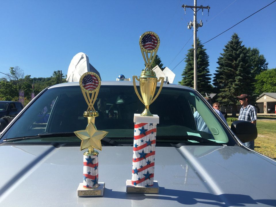 Two trophies on the hood of a silver car at an outdoor event.