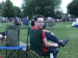 Person sitting in a green folding chair at a grassy park with other people in the background