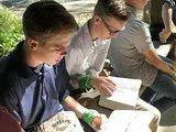 Two people in a classroom read papers at a desk, one holding a green pen