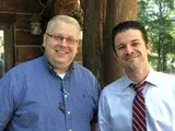 Two smiling men standing outdoors, one in a blue shirt and one in a white shirt with a red striped tie.