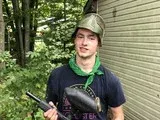 Man holding a handgun, standing outdoors beside a corrugated wall in a wooded area, wearing a helmet and green bandana