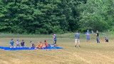 People sitting on blue mats in a grassy field while others stand nearby under a cloudy sky