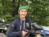 Smiling person in a green cap leaning on a blue motorcycle outdoors among trees
