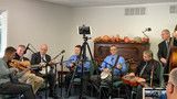 Musicians rehearsing in a small room with guitars, drums, and microphones around a central camera tripod.