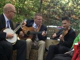 Three musicians seated outdoors playing banjo and violins in a garden setting