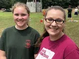 Two smiling people outdoors, one in a green shirt and one in a pink shirt, standing on grass.