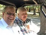 Two smiling men sitting outdoors in the shade, one taking a selfie