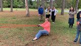 Person in pink sits on grass pulling a rope while others stand nearby in a park.