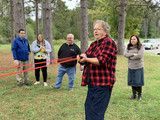 Woman cutting a red ribbon at an outdoor ceremony while three people stand nearby in a wooded area