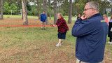 People standing outdoors in a park, one using a camera, with trees and grass in the background